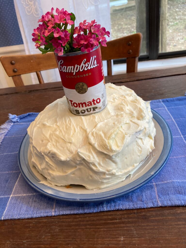 two layer cake with white frosting decorated with a tomato soup can as a vase