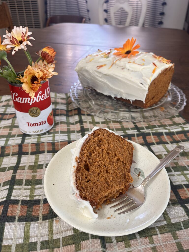 table set with a slice of spice cake topped with white frosting and a can of tomato soup nearby. Cake is in the background.